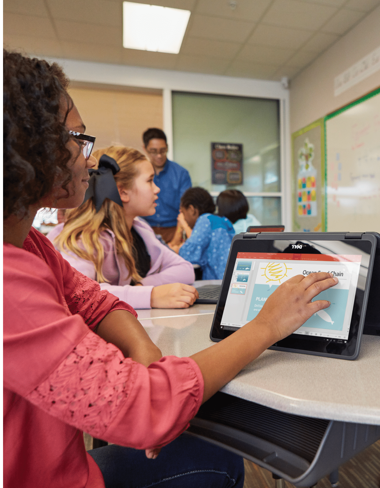 Student seated at desk using a Dell Latitude 11 3000 Series (Model 3189, Keystone 11) notebook computer, while two students are presenting at the front of the classroom  
