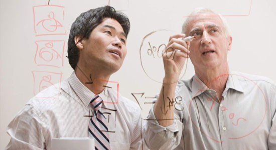 Two men stand at a clear whiteboard drawing diagrams