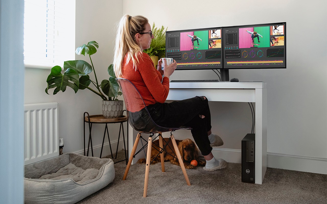 Woman holds coffee as she works on Dell Optiplex system and monitors, with dog under her desk at home.