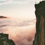 Person standing on cliff edge above low level of clouds.