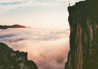 Person standing on cliff edge above low level of clouds.