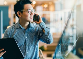 Young confident man strides through office, holding a folder and talking on a cell phone.