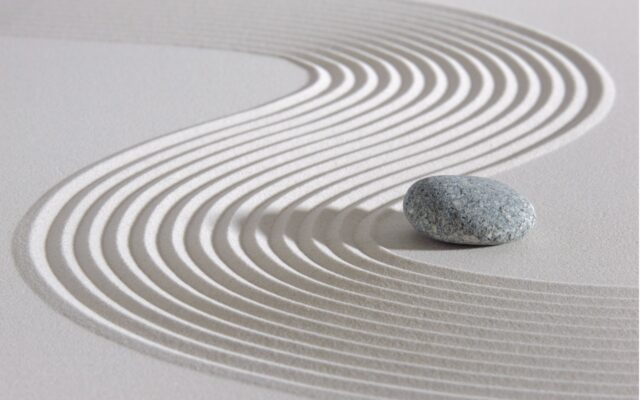 Japanese Zen garden close up of white sand raked into a curve pattern around a small gray stone.