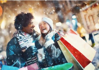 Man and woman with shopping bags smiling in the snow with light from behind.