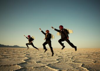 Three people dressed in suits with airplane wings strapped to their backs jump up in the desert.