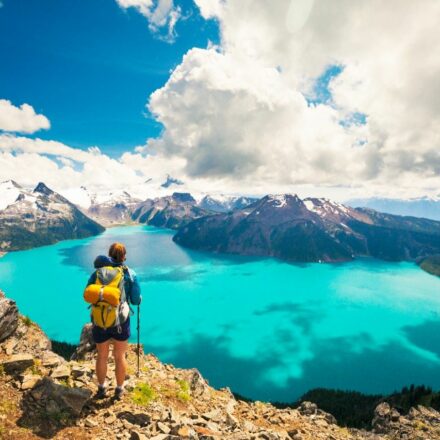 Backpacker stands on a cliff overlooking a blue lake and a mountains in the distance.