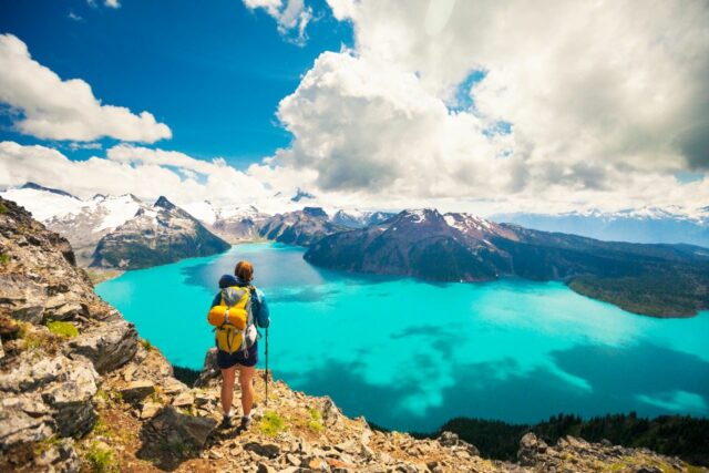 Backpacker stands on a cliff overlooking a blue lake and a mountains in the distance.