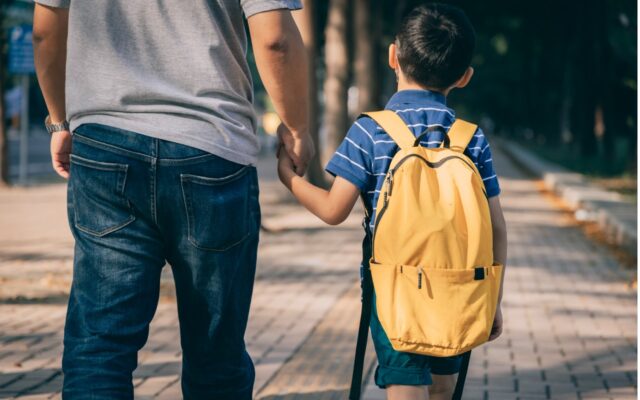 Father walking his son to kindergarten, walking away from camera. Boy has a yellow backpack.