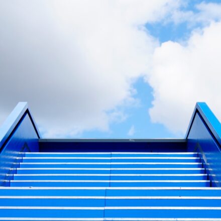 Blue colored flight of stairs leading up towards's the sky with some clouds overhead.
