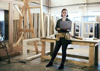 Woman stand in a carpentry shop, wearing a tool belt and holding a Dell computer.