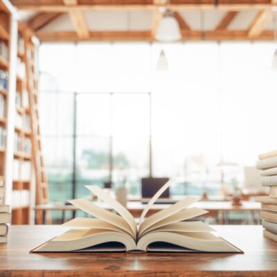 An open book on a table between two stacks of books, and library window in the background.