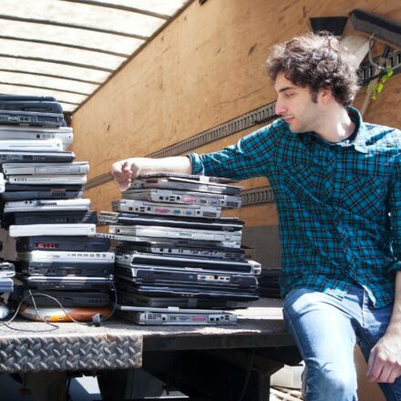 Man in green plaid shirt sits on back of cargo truck next to stack of old laptop computers to be recycled.