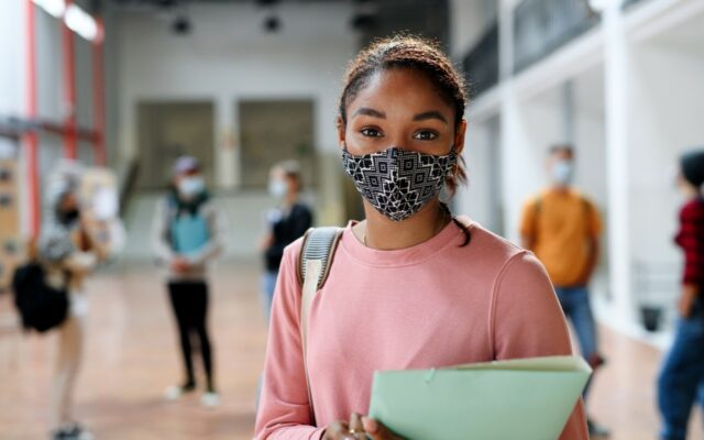 Young African-American female student at school, wearing facial covering as other students in the background also wear facial coverings.
