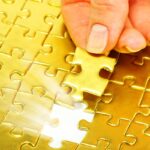 Woman holding gold color puzzle piece over spot in puzzle where it fits.