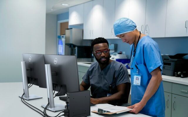 Male and female medical professionals confer about patient case with Dell Optiplex 3000 Thin Client and P2422H monitors on desk in front of them.