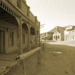 Image of Old West American town, dusty street, hitching post and wooden buildings, in sepia tone.