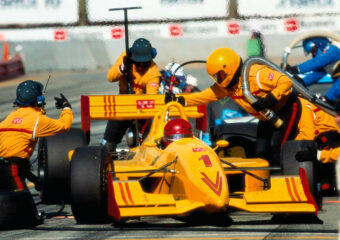 F1 race car team in yellow outfits work on the yellow race car with red stylized design during a pit stop.