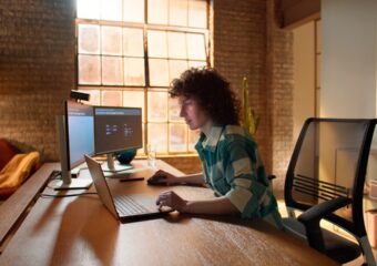 Person in green and white checkered shirt works remotely using a Dell Precision 5000 series model laptop and dual monitors.