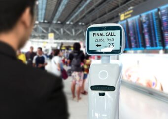 View over a traveller's shoulder of robotic assistance providing flight and gate information in an airport terminal.