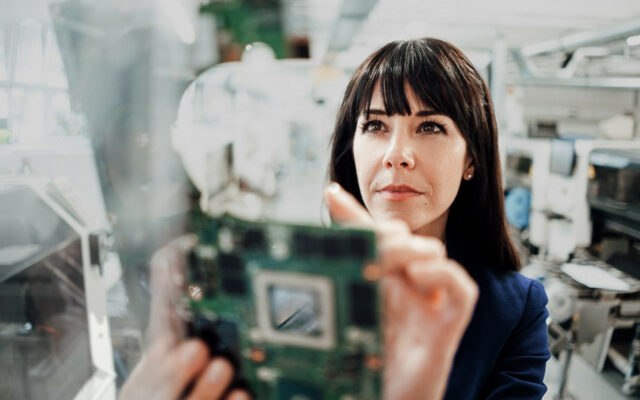 Woman holding up circuit board component in innovation development facility.