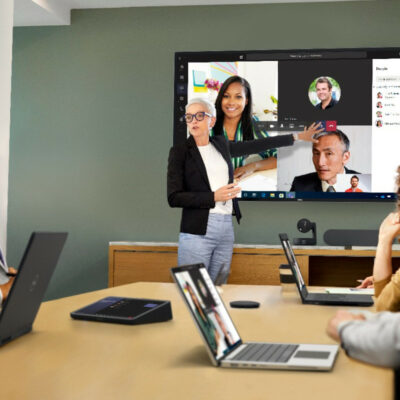 Woman leads presentation in office meeting room in front of large display showing colleagues who are taking part in the meeting from remote locations.