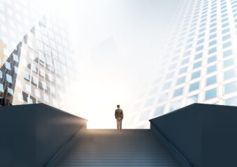 Angle from below of a businessperson at the top of a long flight of outdoor stairs, between two high rise buildings. Representative of reaching a milestone.