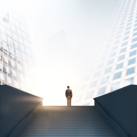 Angle from below of a businessperson at the top of a long flight of outdoor stairs, between two high rise buildings. Representative of reaching a milestone.