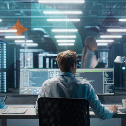 Photo from behind a man working at dual monitor workstation with coding on screen. In the background a woman walks in data center area, behind glass. Textured pattern overlay can be seen with orange diamond shape near upper left corner.