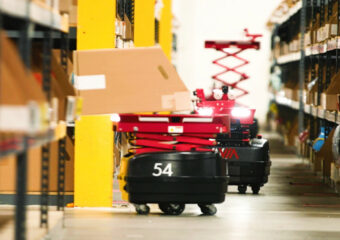 Two InVia Robotics robots, red and black in color, perform warehouse stocking tasks. One robot is in the background with a vertical extender to load higher level racks. Robot number 54 is in the foreground loading a box onto a warehouse shelf.
