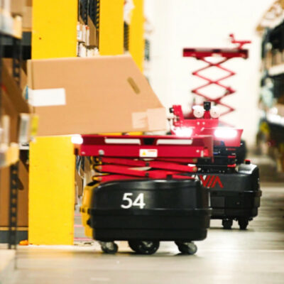 Two InVia Robotics robots, red and black in color, perform warehouse stocking tasks. One robot is in the background with a vertical extender to load higher level racks. Robot number 54 is in the foreground loading a box onto a warehouse shelf.