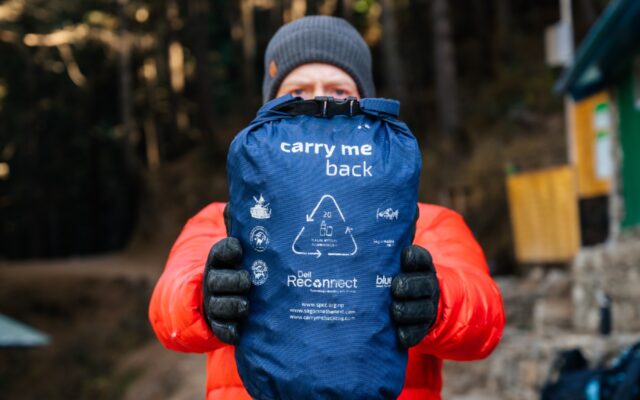 Man in gray ski cap, orange winter jacket, holds a Carry Me Back initiative pack, to bring back trash a traveler's trash in Nepal's Sagarmatha National Park and the Khumbu region. Photo credit by Martin Edstrom