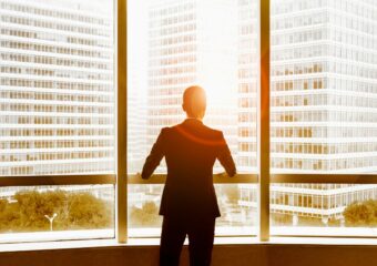 View from behind of a businessman looking out office windows at nearby buildings and sunlight emerging between buildings.
