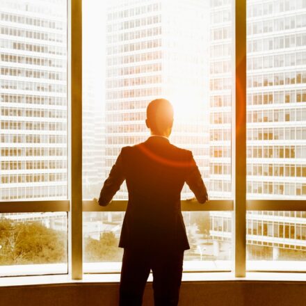 View from behind of a businessman looking out office windows at nearby buildings and sunlight emerging between buildings.