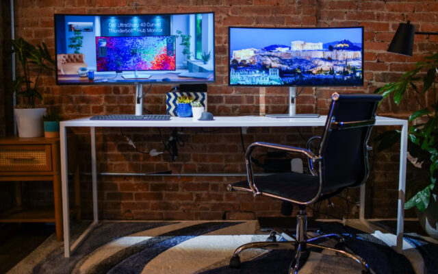 Dell Ultra Sharp 40" E 34" Monitor affiancati su una scrivania di fronte a un muro di mattoni con una sedia da ufficio di fronte.